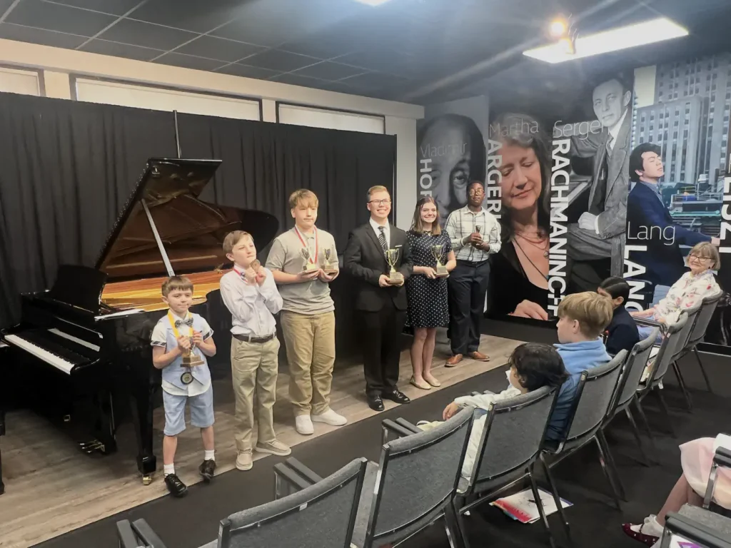 Young piano competition winners holding trophies on stage