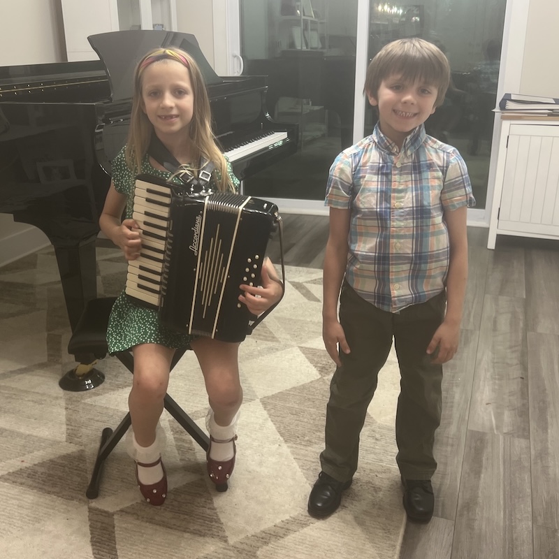 Young girl playing accordion with smiling boy beside her