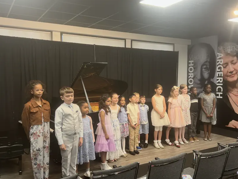 Children lined up on stage near a grand piano recital